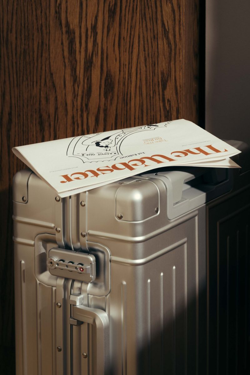 A silver hard-shell suitcase stands upright next to a wooden door, with a folded newspaper from The Webster partially covering its handle and top, featuring bold orange text and a graphic illustration on the front page.