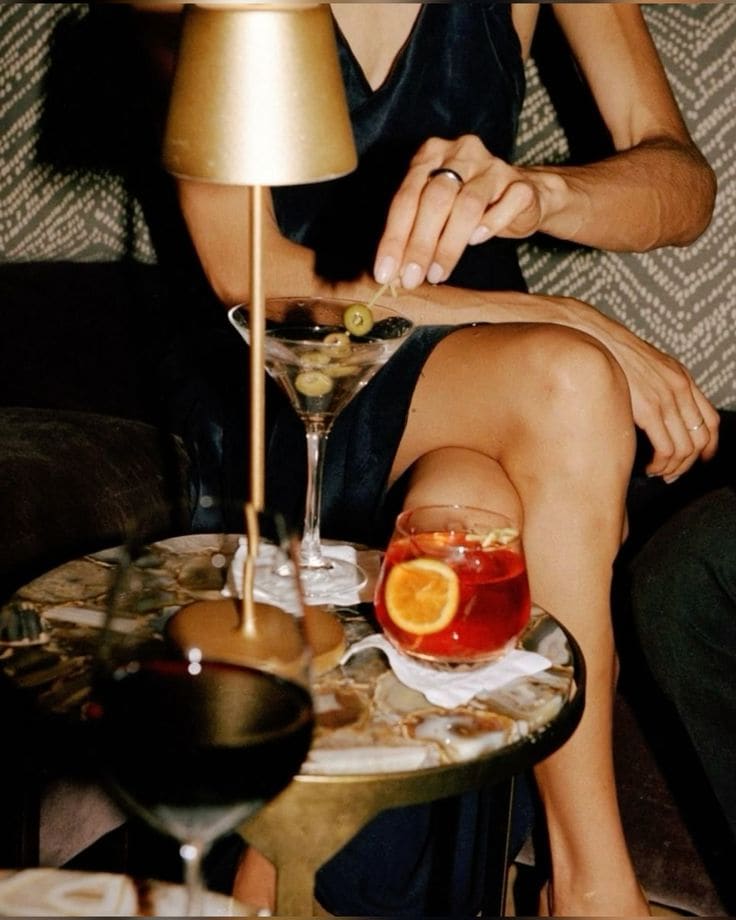 A person in a dark dress sits at a small table in Covent Garden, adjusting an olive in a martini glass. The table holds a red cocktail with an orange slice, a glass of red wine, and a gold lamp against the dimly lit background.