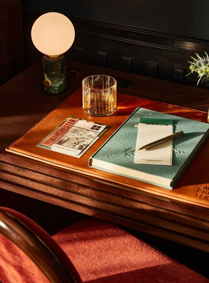A cozy wooden desk with a lit round lamp, a glass of amber drink, and a light green notebook evokes the inviting charm of Covent Garden. Warm, soft lighting enhances the scene atop a leather desk pad with notepad and magazine.