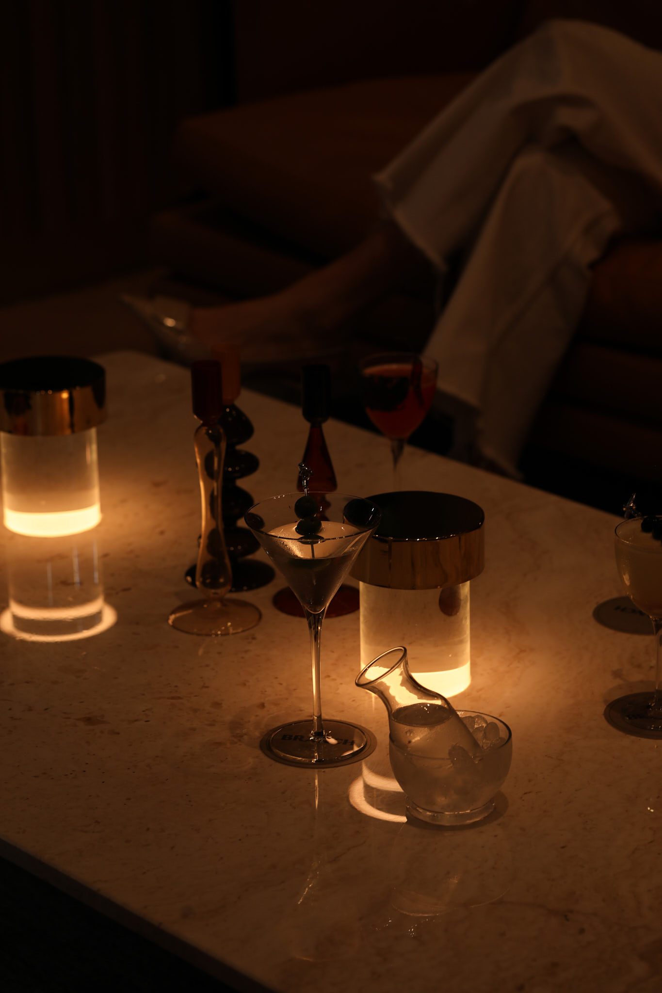 A dimly lit table in Covent Garden with glowing candles, a martini glass with olives, a small carafe with ice, and assorted bottles. In the background, a person in white pants is seated on a couch.