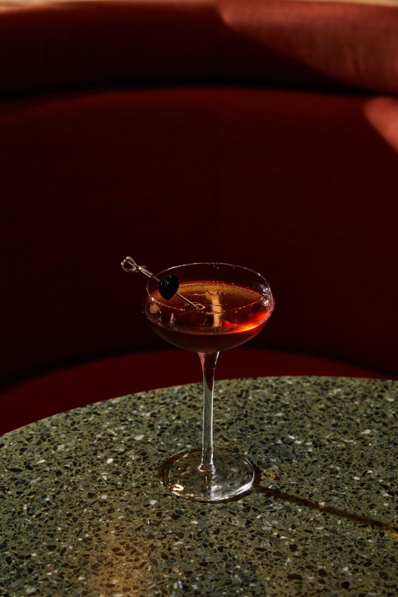 A cocktail in a coupe glass with a cherry garnish rests on a speckled green tabletop in Covent Garden, with a curved red upholstered booth in the background.