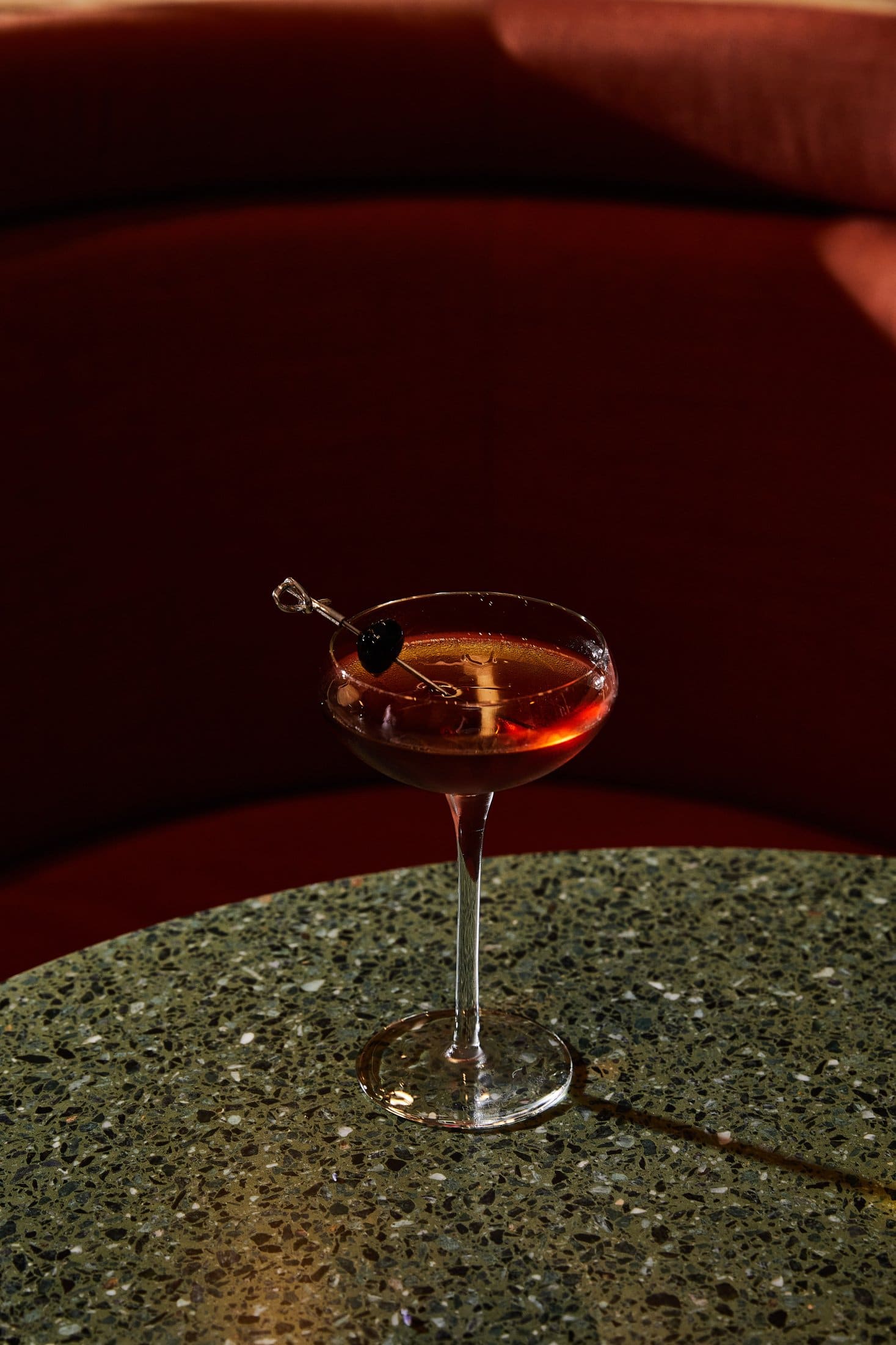 A cocktail in a coupe glass with a cherry garnish rests on a speckled green tabletop in Covent Garden, with a curved red upholstered booth in the background.