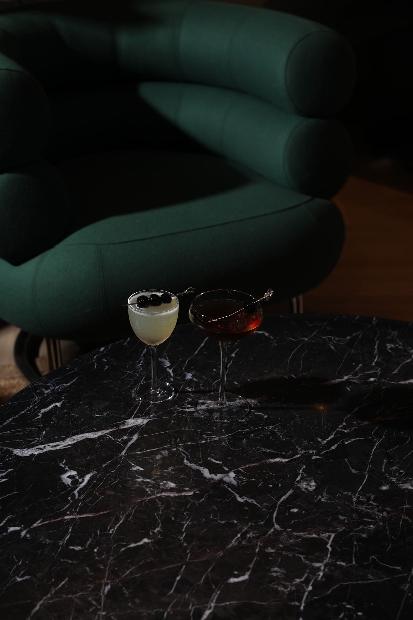 Two elegant cocktails, one light with berries and one dark with a cherry garnish, sit on a black marble table in front of a plush, dark green chair—perfect for an evening in stylish Covent Garden.