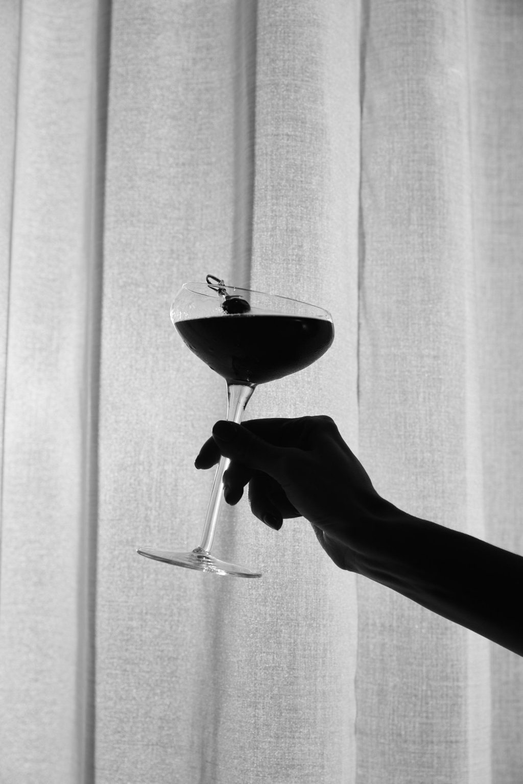 A hand holds up a cocktail glass filled with a dark drink and garnished with a cherry, silhouetted against light curtains—capturing the elegant vibe of Covent Garden nightlife.