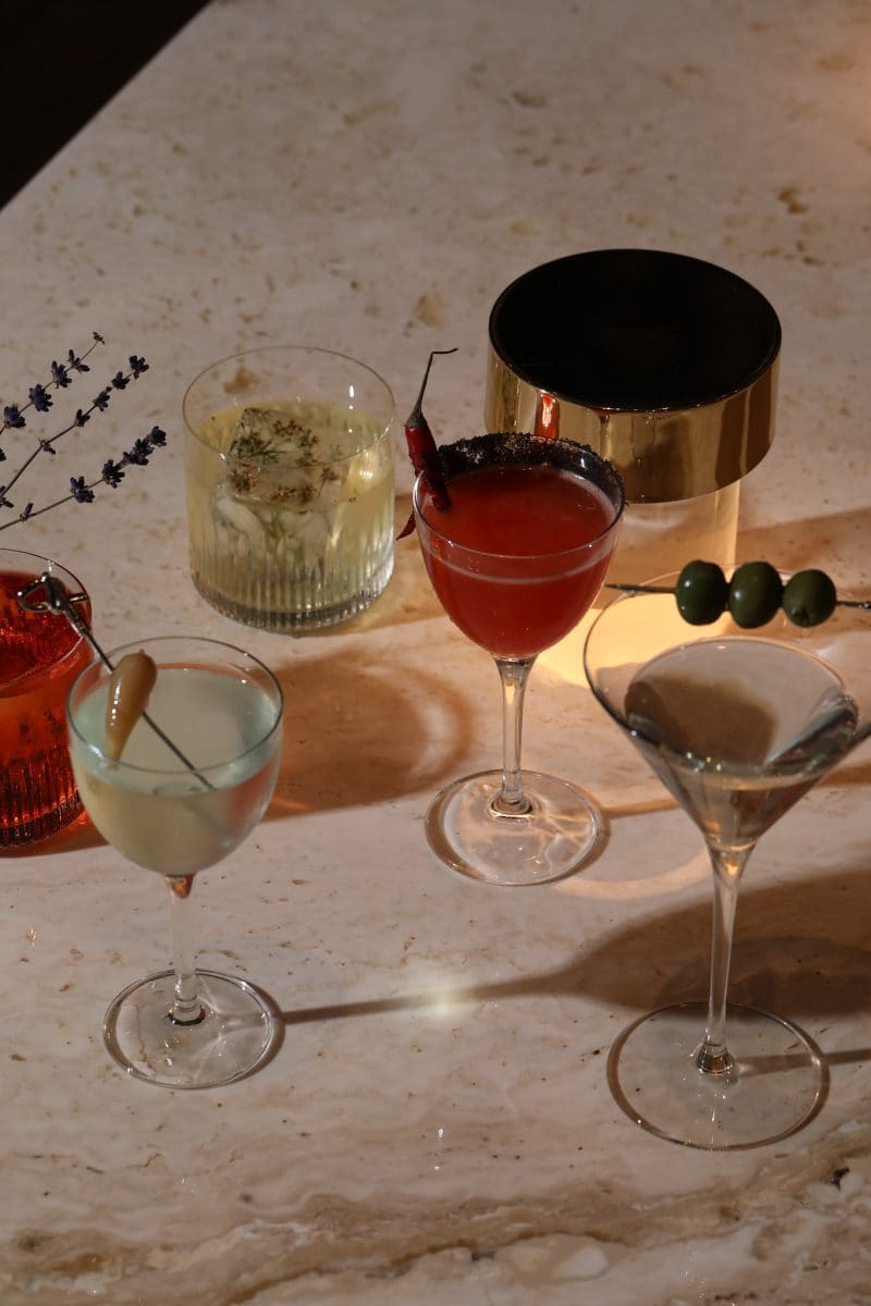 Five assorted cocktails in elegant glasses sit on a marble table in Covent Garden, each garnished uniquely with herbs, peppers, olives, flowers, or fruit. Warm lighting highlights their vibrant colors and stylish presentation.