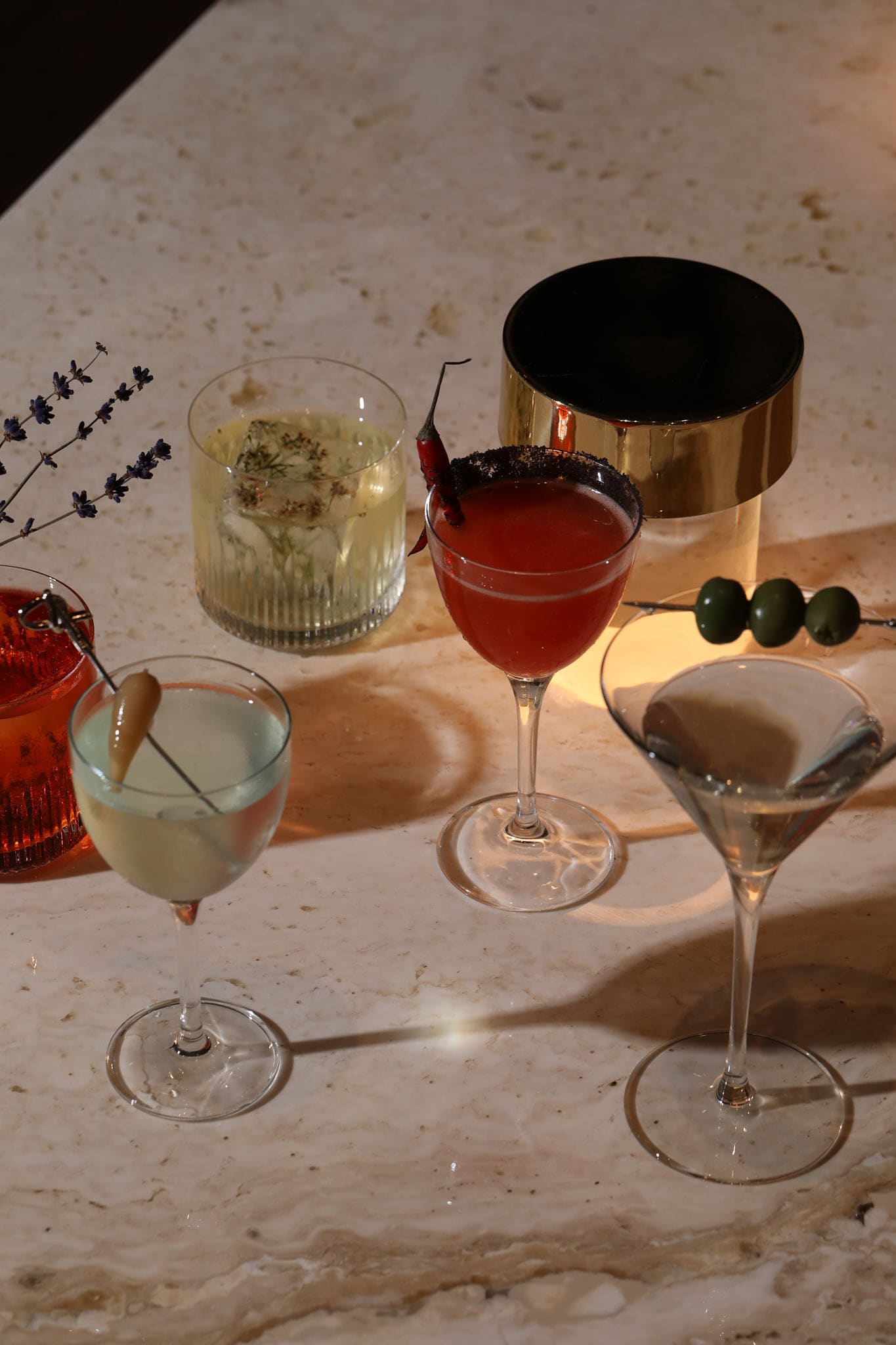 Five assorted cocktails in elegant glasses sit on a marble table in Covent Garden, each garnished uniquely with herbs, peppers, olives, flowers, or fruit. Warm lighting highlights their vibrant colors and stylish presentation.
