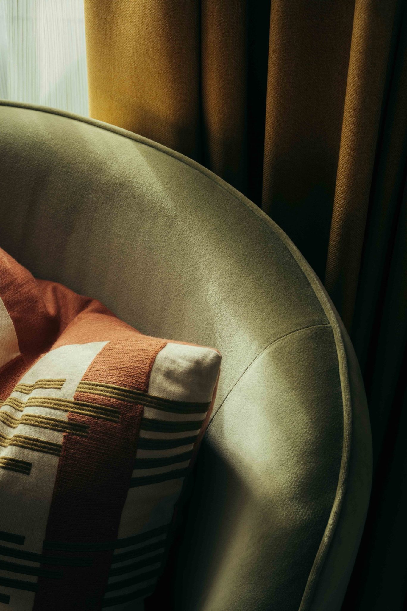 A close-up of a beige velvet armchair with a geometric-patterned cushion in warm tones, next to mustard yellow curtains in a sunlit Covent Garden-inspired setting.