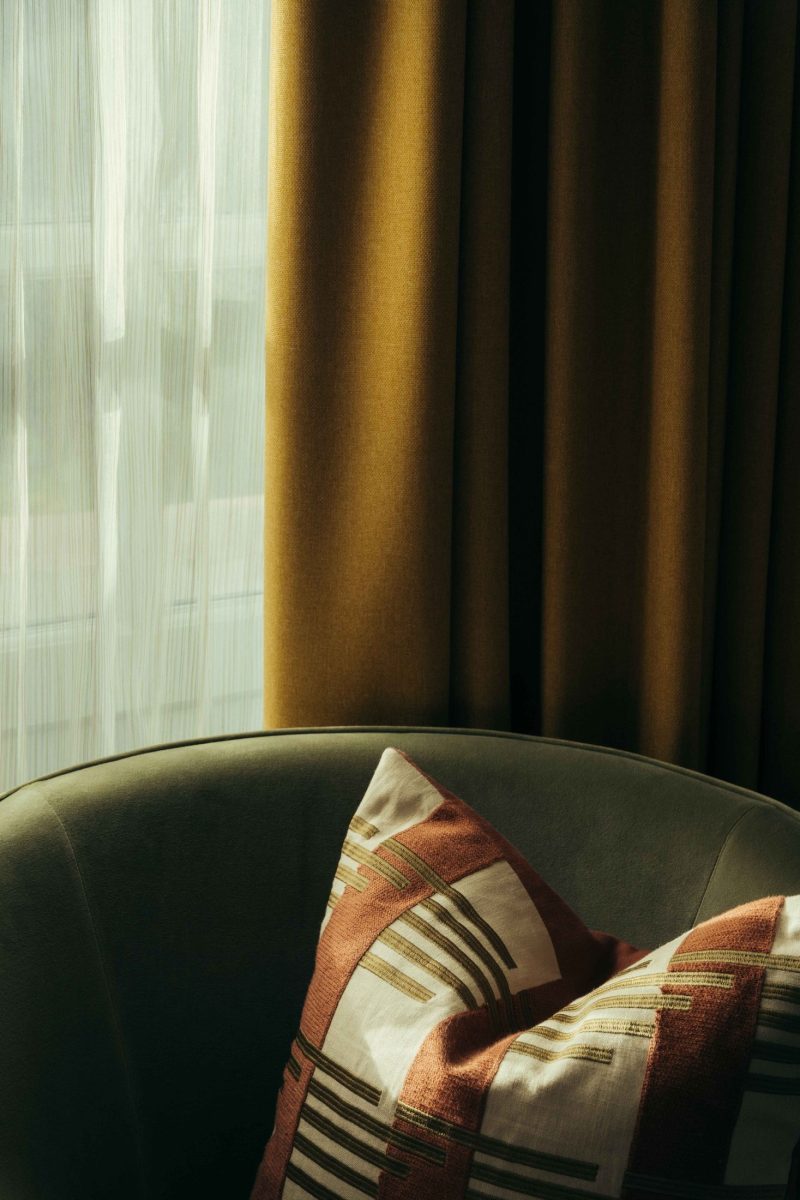 A soft, green armchair with a geometric-patterned pillow sits beside a window with sheer white curtains and mustard yellow drapes, evoking the relaxed charm of Covent Garden, all softly lit by natural daylight.