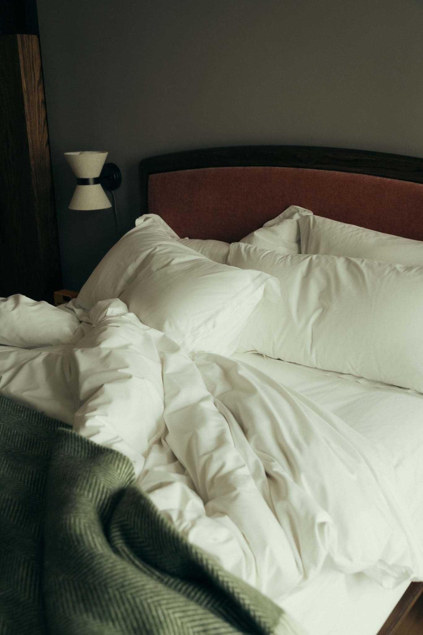 A neatly made bed with white pillows and comforter, a wooden headboard, and a green patterned blanket at the foot. Reminiscent of Covent Garden charm, a wall-mounted lamp glows on the left side against a gray wall.