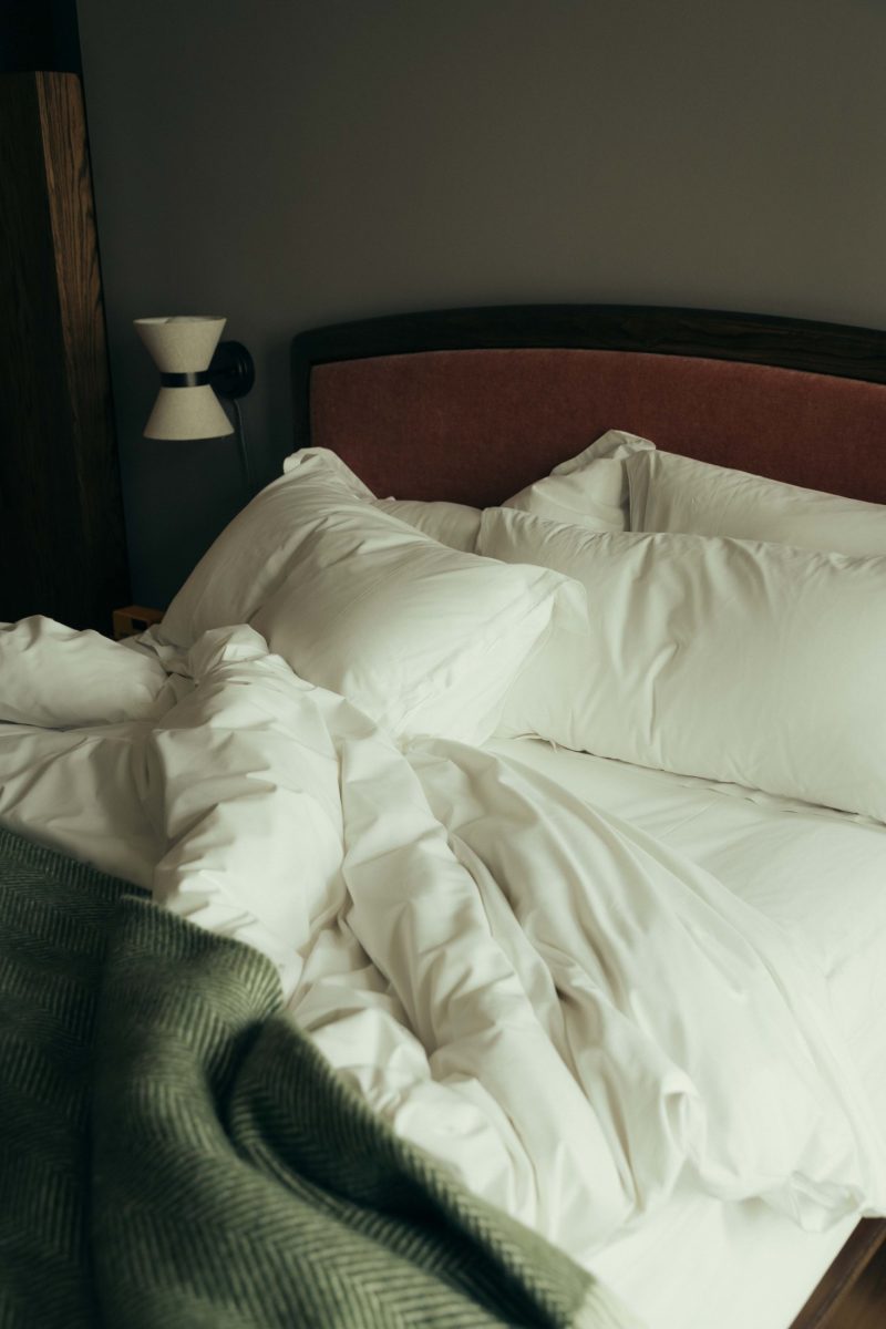 A neatly made bed with white pillows and comforter, a wooden headboard, and a green patterned blanket at the foot. Reminiscent of Covent Garden charm, a wall-mounted lamp glows on the left side against a gray wall.