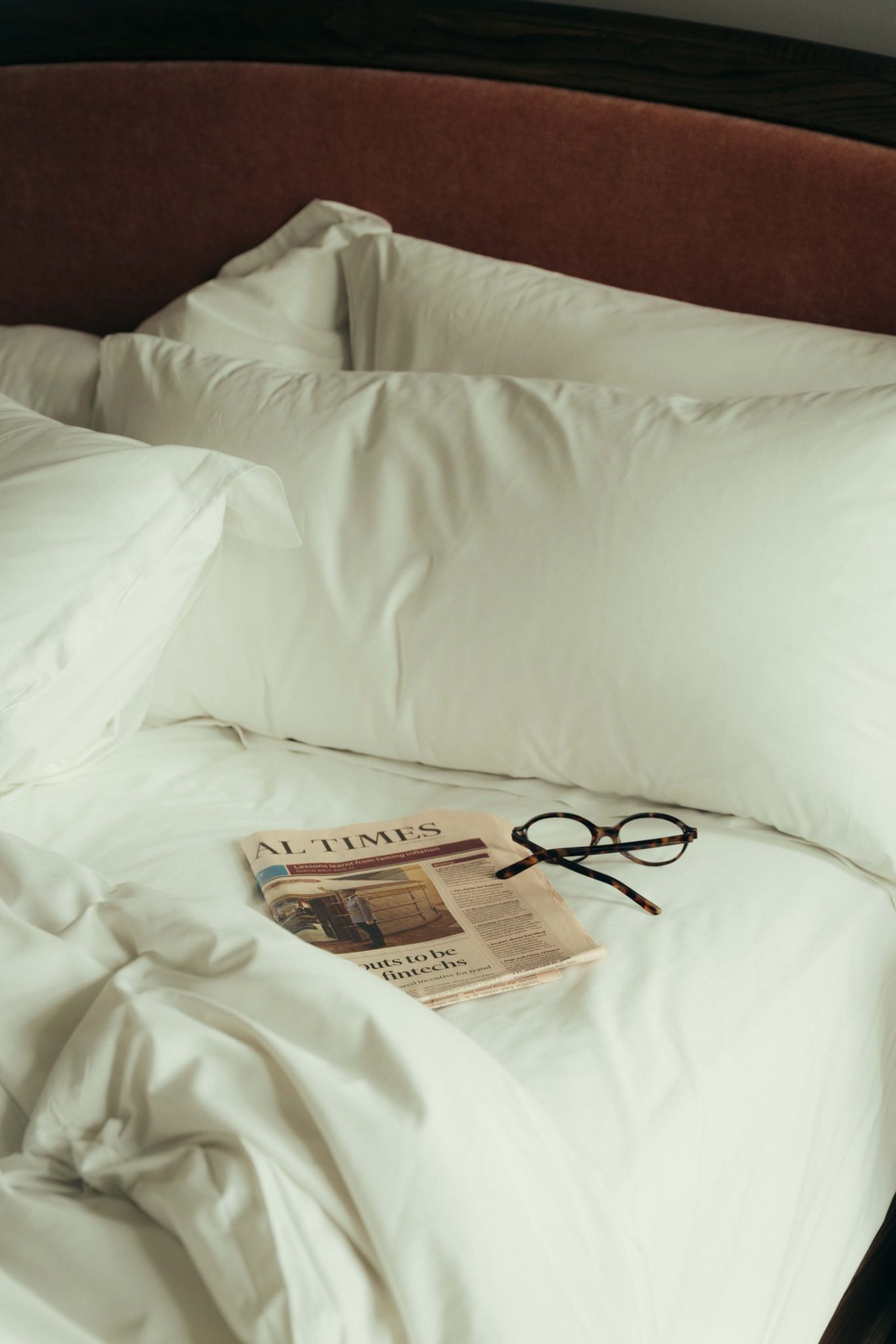 A neatly made bed with white sheets and pillows has a folded newspaper and a pair of black eyeglasses resting on top, evoking the cozy charm of a Covent Garden morning.
