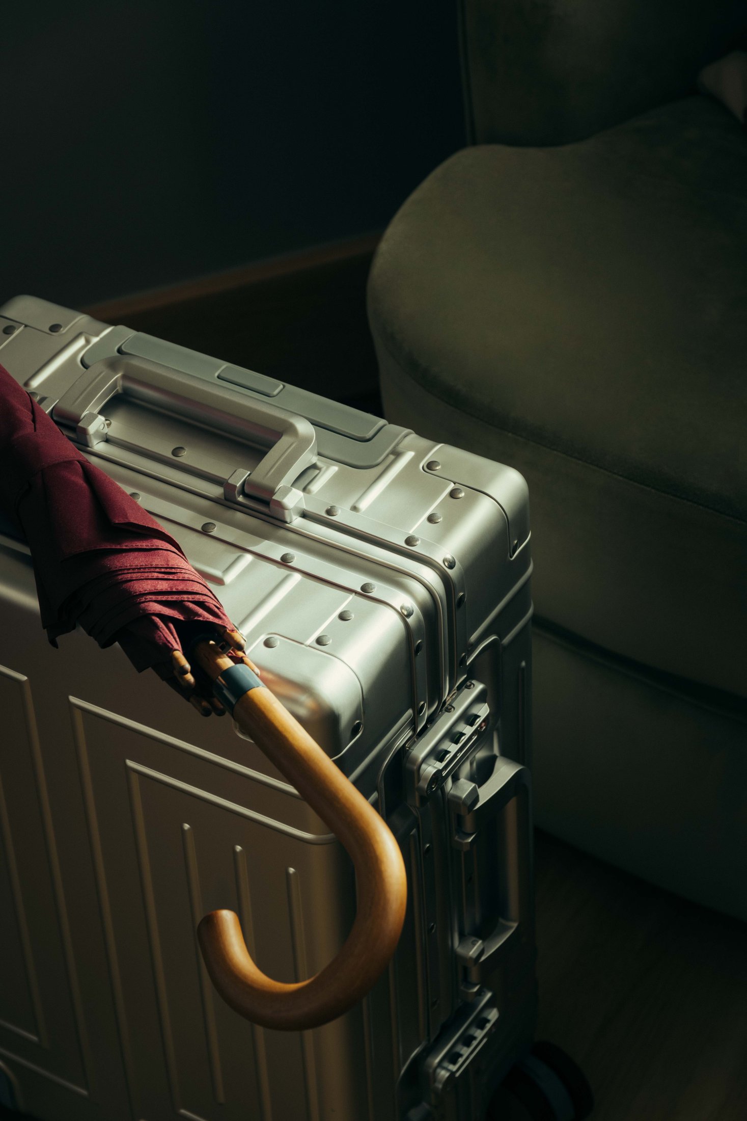 A silver hard-shell suitcase stands next to a green armchair, with a burgundy umbrella featuring a wooden curved handle resting on top—evoking the timeless charm of Covent Garden. Soft light highlights the suitcase and umbrella.