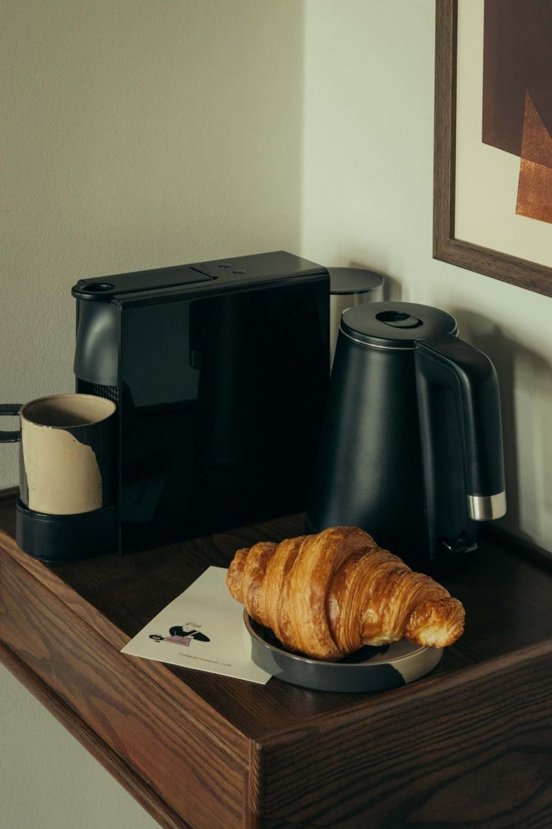 A black coffee maker, black electric kettle, and a mug sit on a wooden shelf. In the foreground is a plate with a croissant and a piece of paper, evoking the cozy charm of Covent Garden, next to a framed artwork on the wall.