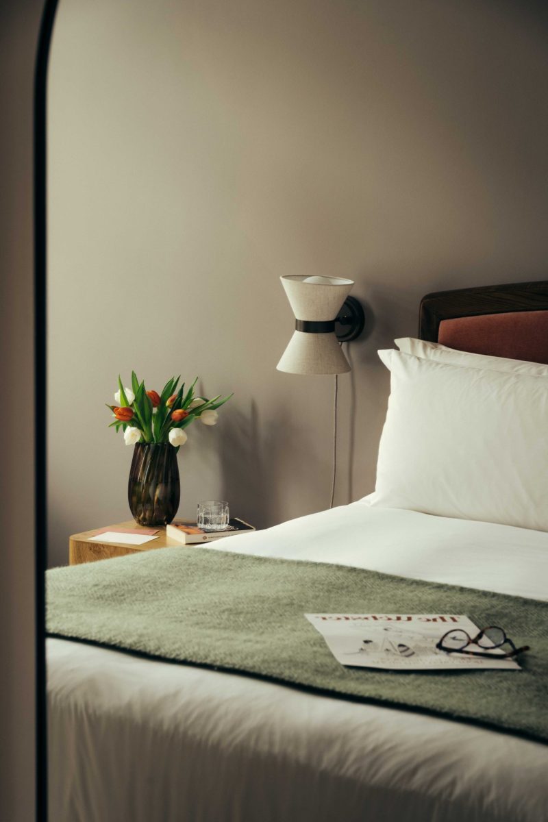 A neatly made bed with white linens and a green throw blanket sits beside a wooden nightstand holding a vase of tulips, a glass of water, and a wall-mounted lamp; perfect for relaxing after exploring Covent Garden, with a magazine and glasses resting on the bed.