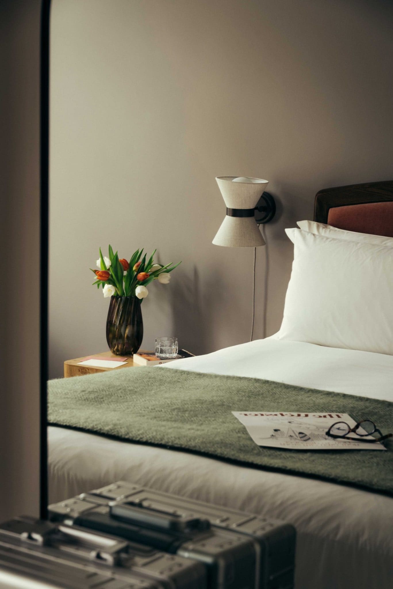 A neatly made bed with white linens and a green throw blanket evokes Covent Garden charm. A nightstand holds tulips, a glass of water, and is lit by a wall lamp. Papers and glasses rest on the bed, with a silver suitcase nearby.