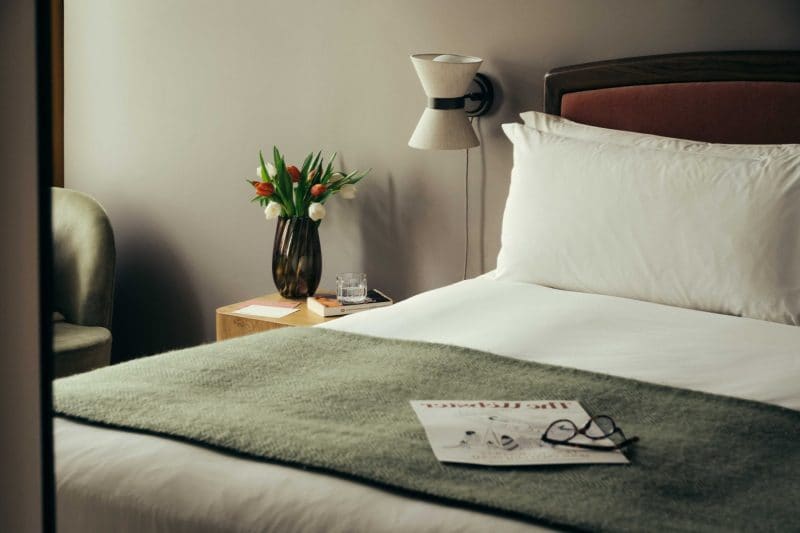 A neatly made bed with white linens and a green throw evokes Covent Garden charm, set beside a table with tulips, a glass of water, and a book. Glasses and a magazine rest invitingly on the bed.