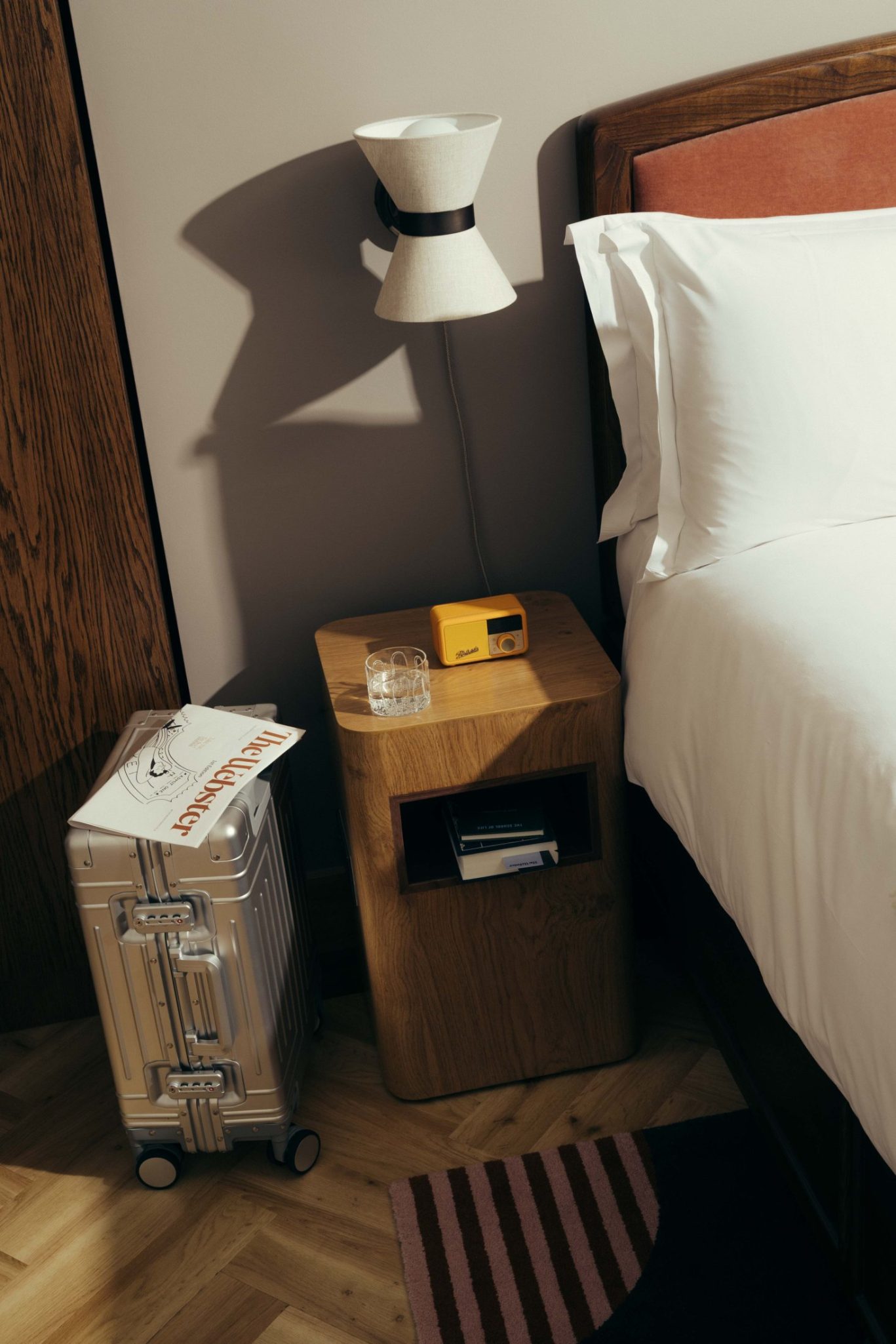A neatly made bed with white sheets beside a wooden nightstand holding a glass, a yellow camera, and books. A lamp is mounted above, and a silver suitcase with papers rests on the floor—evoking Covent Garden’s stylish charm.
