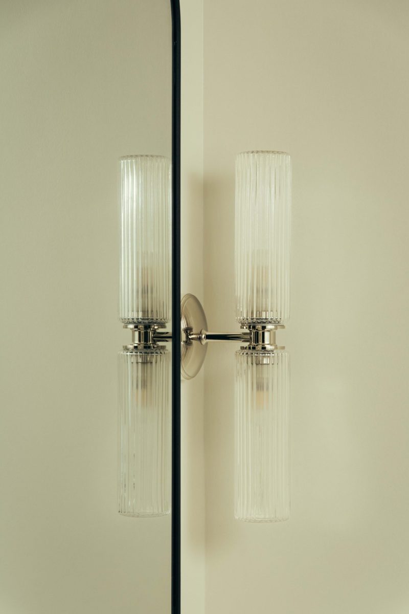 A modern Covent Garden wall sconce with two ribbed glass shades is mounted on a light-colored wall near a mirror that reflects the fixture, creating a symmetrical appearance.