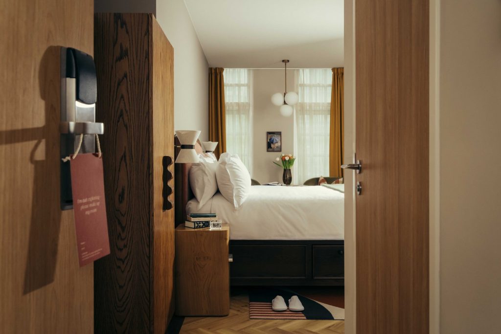 A cozy Covent Garden hotel room with a neatly made bed, bedside tables, and a vase of flowers. Warm light streams through large windows with yellow curtains. Slippers rest on a rug by the bed, and the door is ajar.