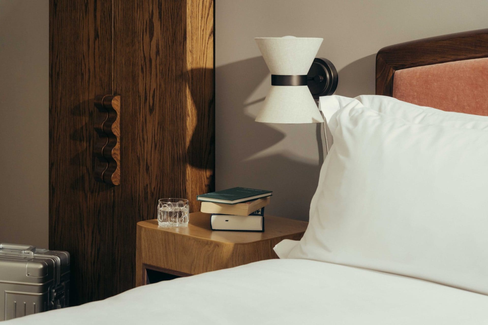 A neatly made bed with white linens beside a wooden nightstand holding a stack of books, a glass of water, and a modern wall lamp, evokes the stylish comfort found in Covent Garden hotels, with a wardrobe and silver suitcase in the background.