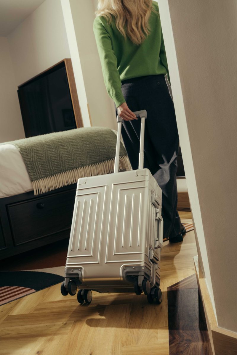A person with long blond hair, wearing a green top and dark pants, pulls a silver rolling suitcase through a modern Covent Garden-style bedroom with a neatly made bed and wooden floor.