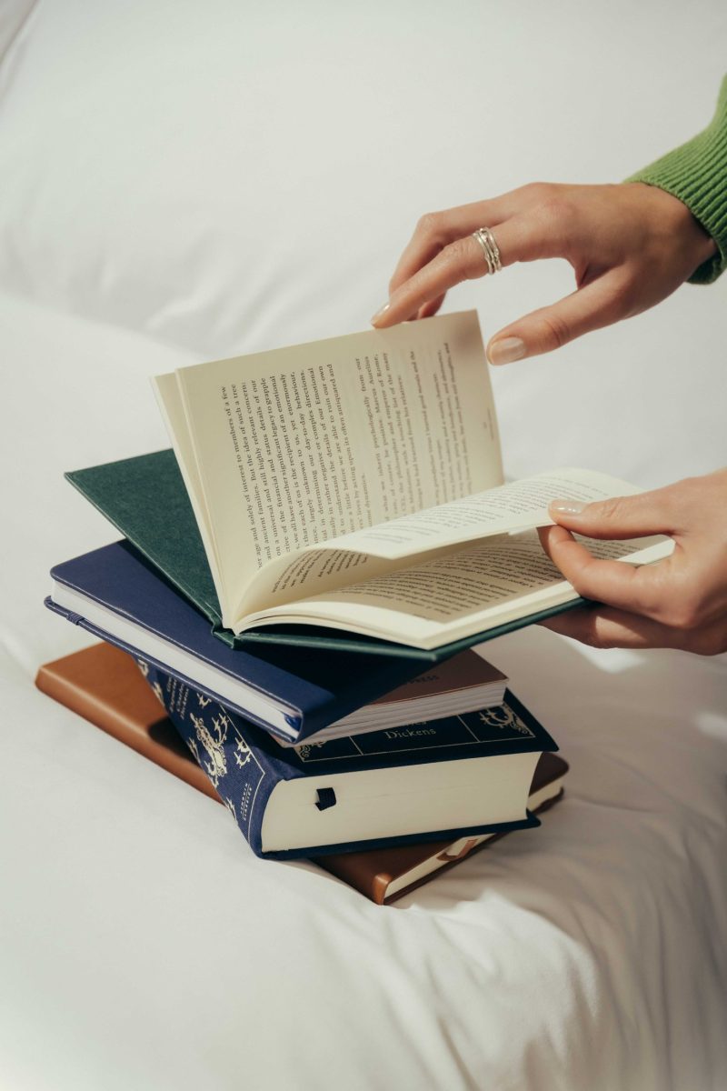 A person with a silver ring and wearing a green sweater flips through an open book, with a stack of closed books resting on a white surface below, reminiscent of a quiet afternoon in Covent Garden.