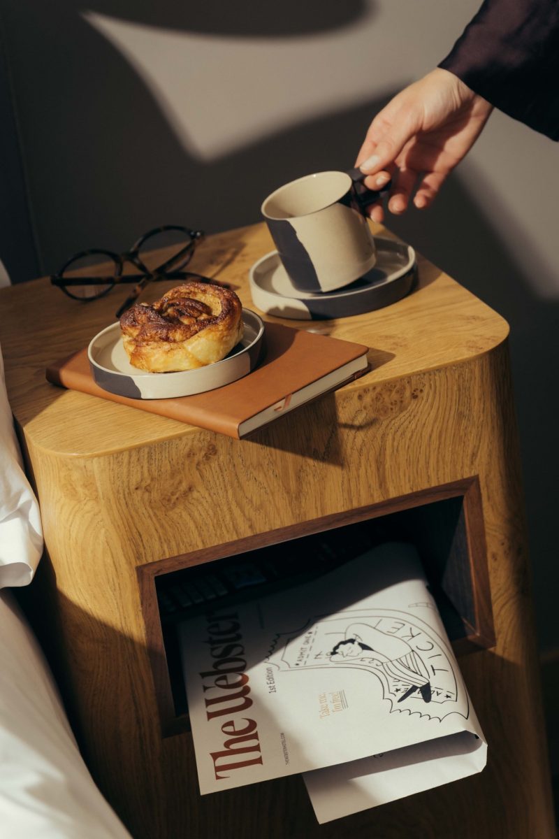 A hand reaches for a ceramic cup on a wooden nightstand, which also holds a saucer with a pastry, eyeglasses, and a notebook—evoking the cozy charm of Covent Garden. A magazine is stored in the shelf below.