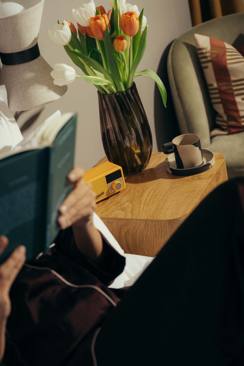 A person reads a book while relaxing indoors. On a wooden side table are a vase of orange and white tulips, a yellow radio, and a cup with a saucer—invoking the cozy charm of Covent Garden. An armchair and hat are in the background.