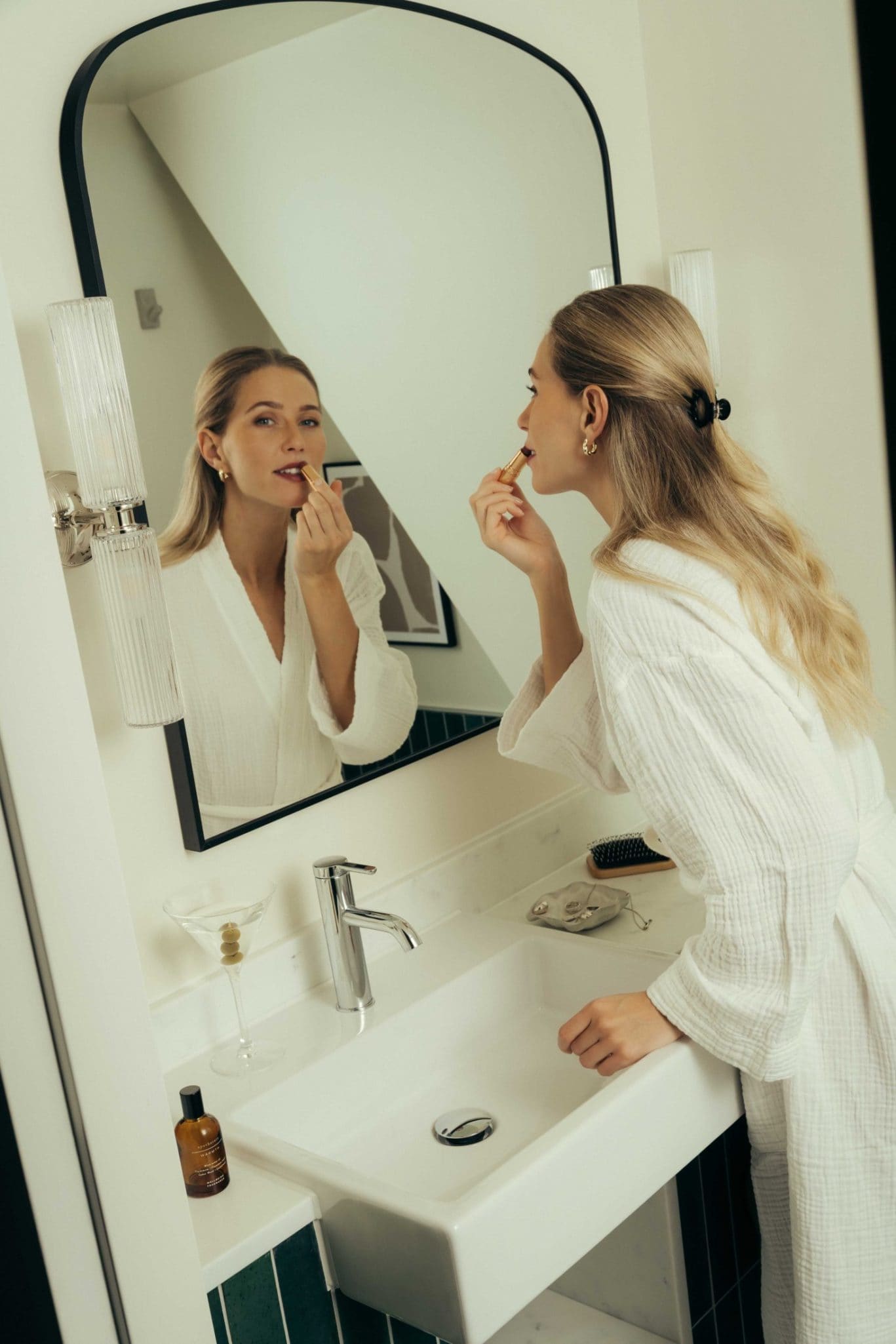 A woman in a white robe applies lipstick while looking in a bathroom mirror, evoking the chic vibe of Covent Garden. Toiletries, a hairbrush, and a martini glass are scattered on the sink counter.
