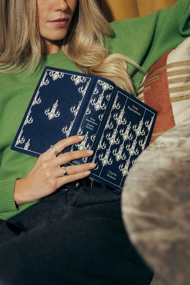 A woman in a green sweater sits on a couch, holding an open navy blue book with chandelier patterns on the cover. The book’s title, Great Expectations by Charles Dickens, is visible—a classic set partially in Covent Garden.