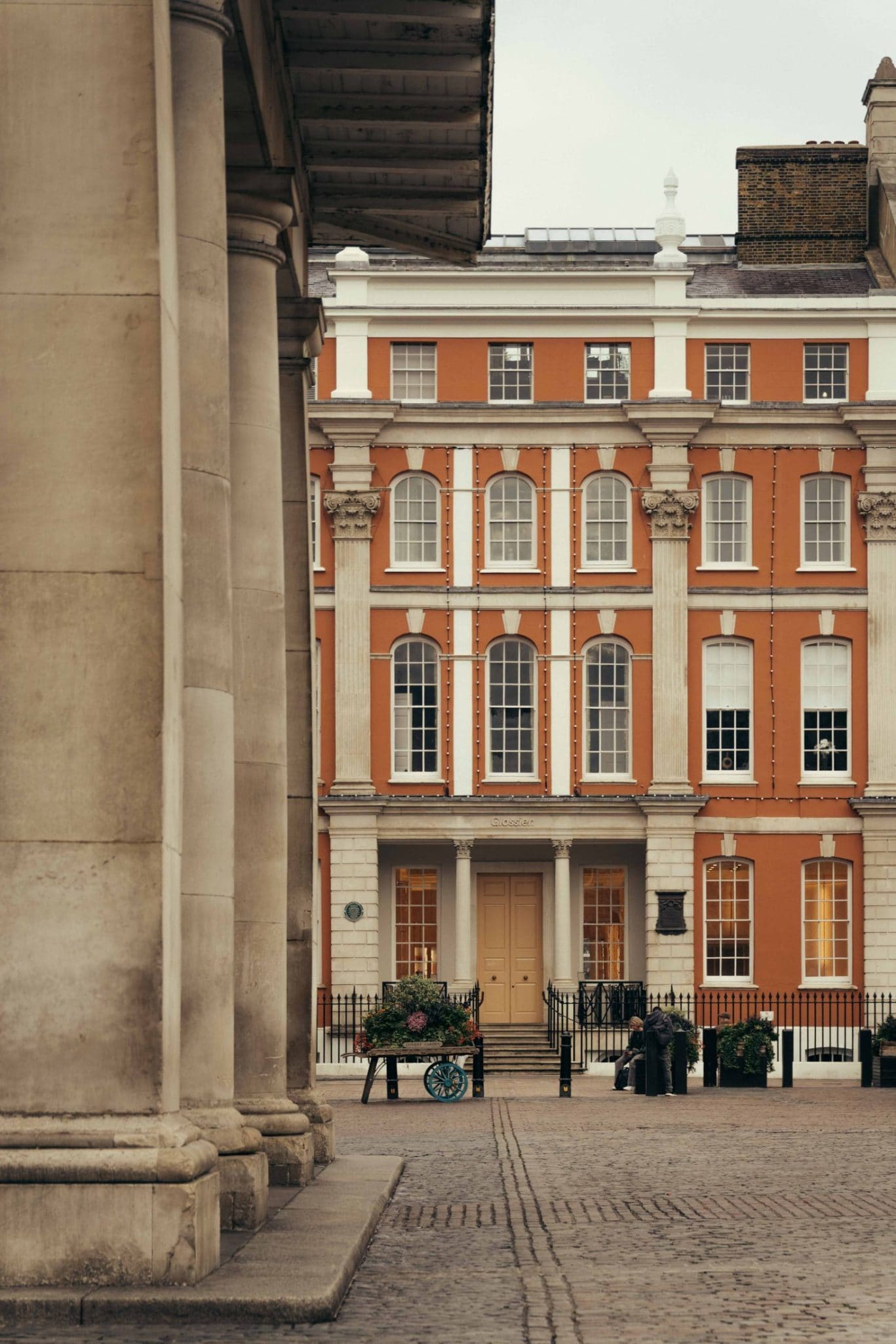A grand orange and white building with tall windows stands behind stone columns in Covent Garden, facing a cobblestone street with a flower cart and two people sitting on a bench.