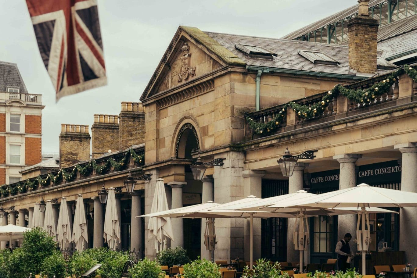 Outdoor seating area with white umbrellas and greenery in front of a historic stone building adorned with garlands in Covent Garden; a British flag is in the foreground, and “Caffè Concerto” signs are visible.