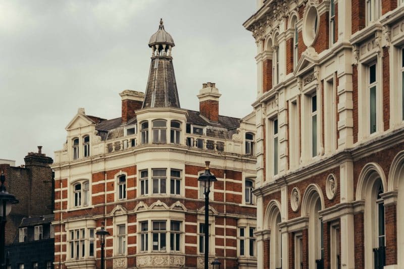 Historic multi-story buildings with ornate facades and red brick details under an overcast sky evoke classic European architecture, reminiscent of Covent Garden’s charming streetscape—one building features a pointed turret and multiple chimneys.