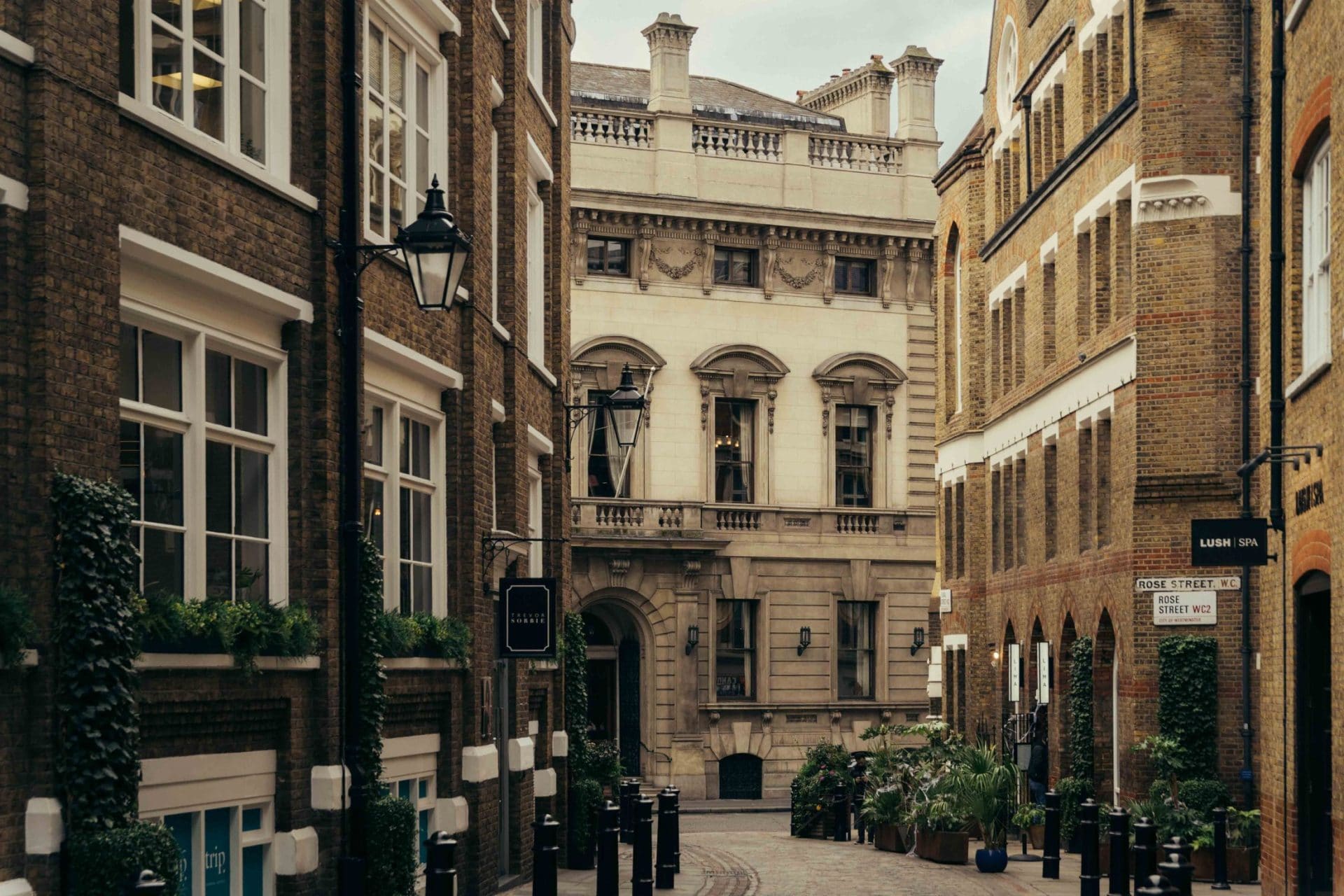 A narrow cobblestone street in Covent Garden, lined with historic brick buildings, street lamps, potted plants, and signs, leads to a grand stone building with tall windows and ornate architectural details.
