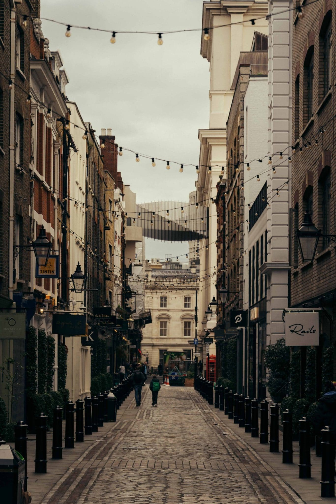 A cobblestone street in Covent Garden is lined with buildings, shops, cafes, and string lights overhead. A few pedestrians walk down the street, and a modern glass bridge connects two buildings in the background.