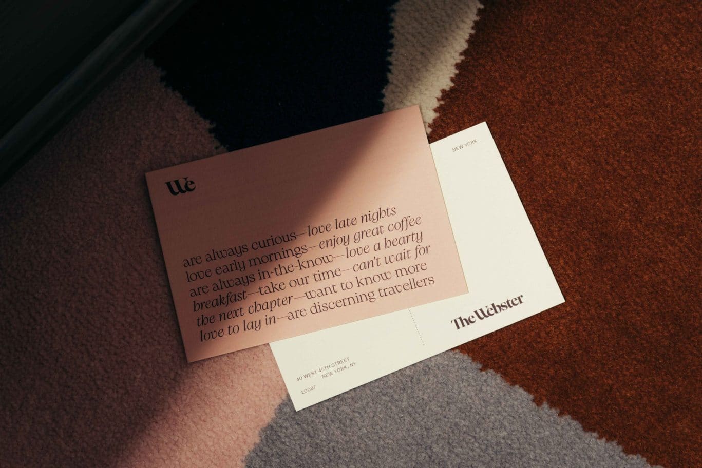 A postcard with poetic text about travel and leisure lies on a multicolored textured carpet. It features phrases about curiosity, late nights, and adventure in Covent Garden, with The Webster printed at the bottom right corner.