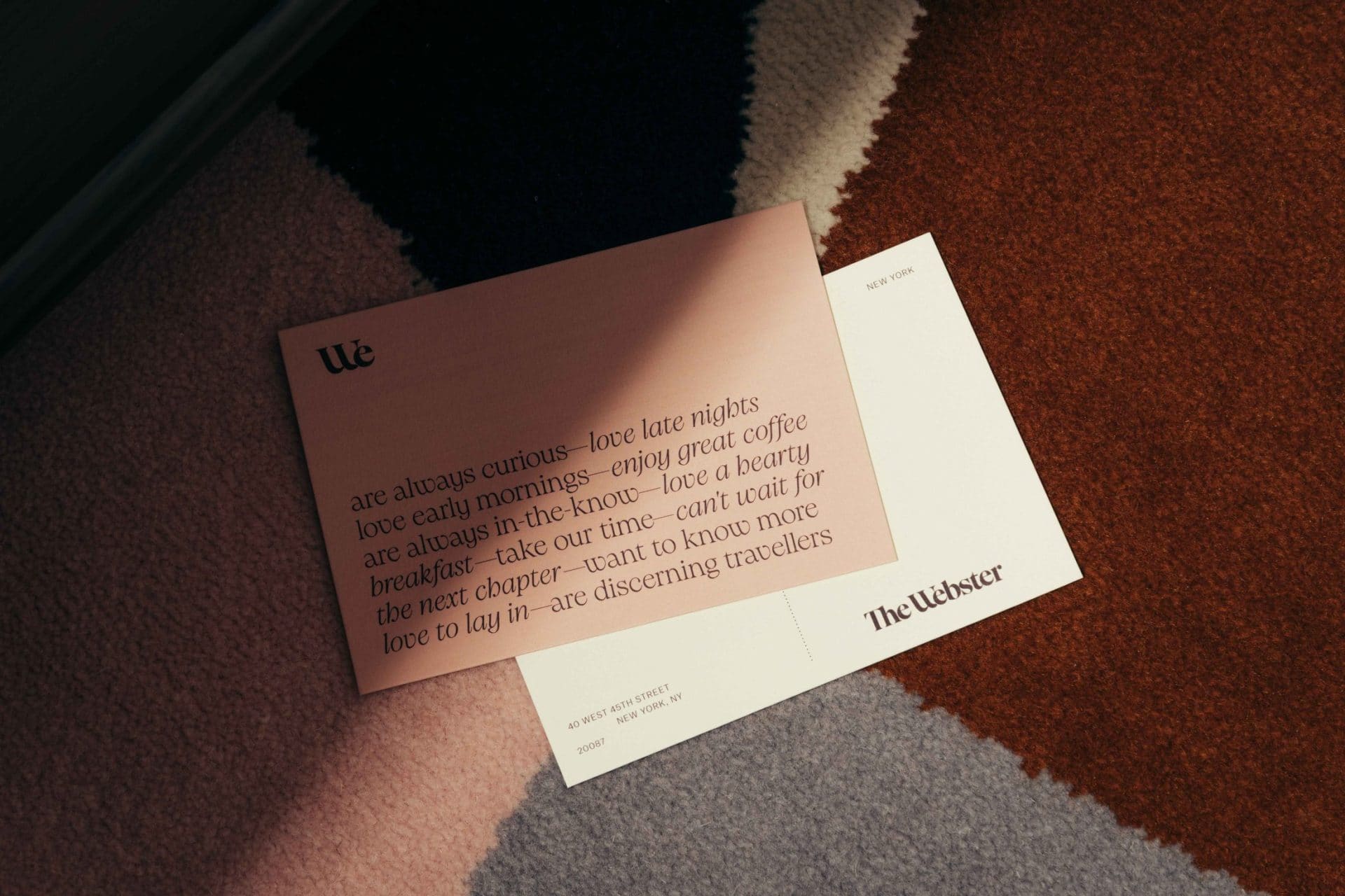 A postcard with poetic text about travel and leisure lies on a multicolored textured carpet. It features phrases about curiosity, late nights, and adventure in Covent Garden, with The Webster printed at the bottom right corner.