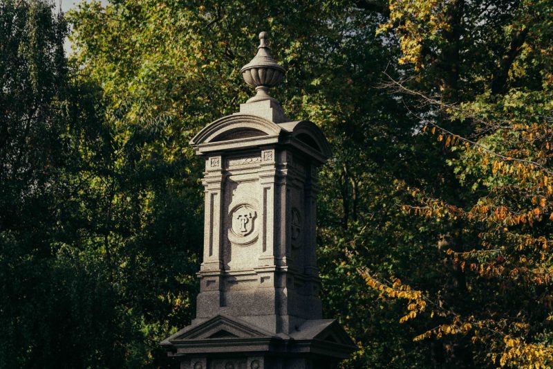 A tall stone monument with decorative carvings and an urn on top stands among green and yellow-leaved trees in a sunlit outdoor setting, reminiscent of the historic charm found near Covent Garden.