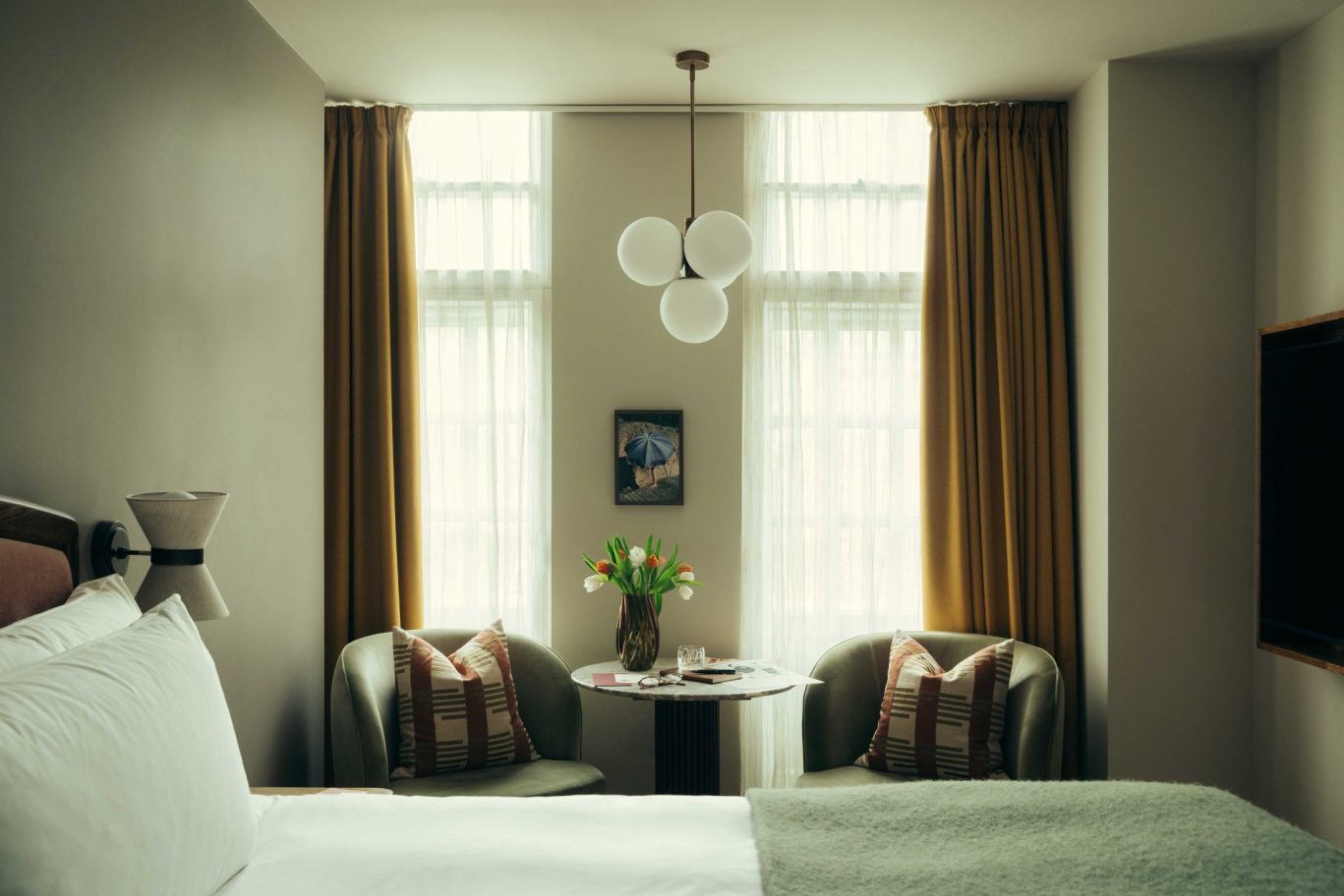 A cozy Covent Garden hotel room with a neatly made bed, two armchairs with patterned pillows by a round table holding a vase of tulips, and two tall windows with mustard curtains letting in soft light.