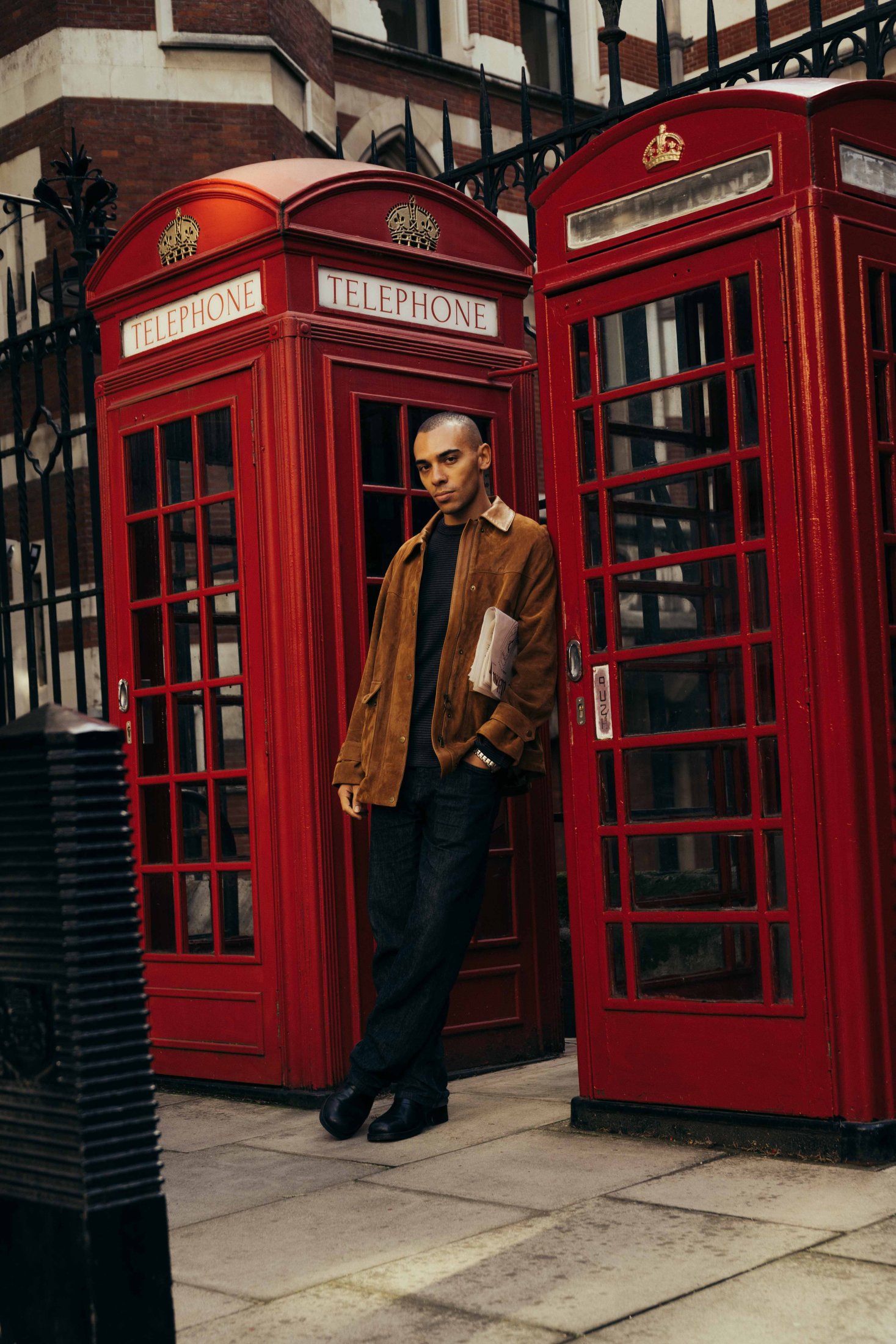 A person leans against a classic red British telephone booth in Covent Garden, holding a book, with another identical booth beside them and a wrought iron fence in the background.