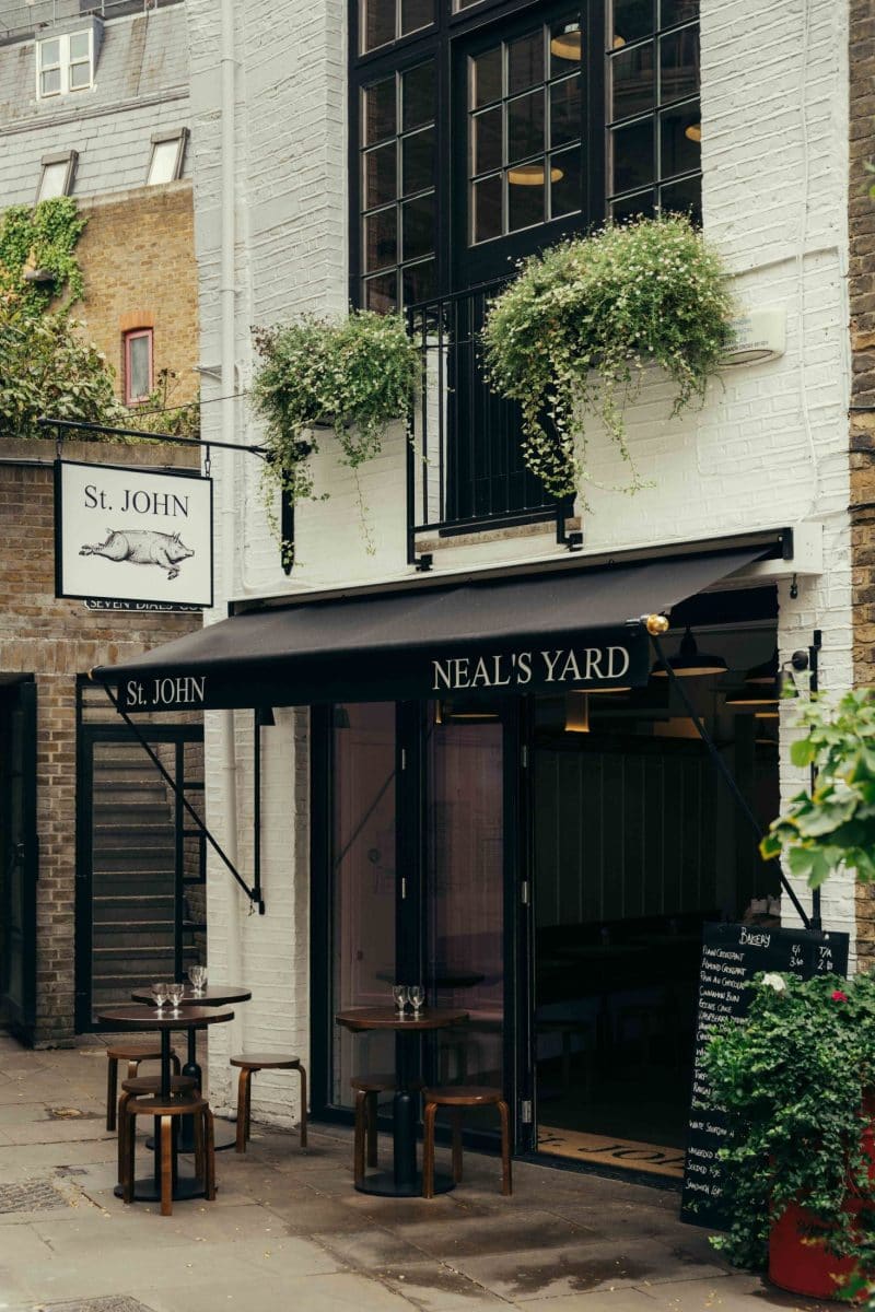 Exterior of St. John restaurant at Neal’s Yard in Covent Garden, featuring a black awning, two small tables with stools outside, potted plants above, a hanging sign, and a menu board near the entrance on a cobblestone street.