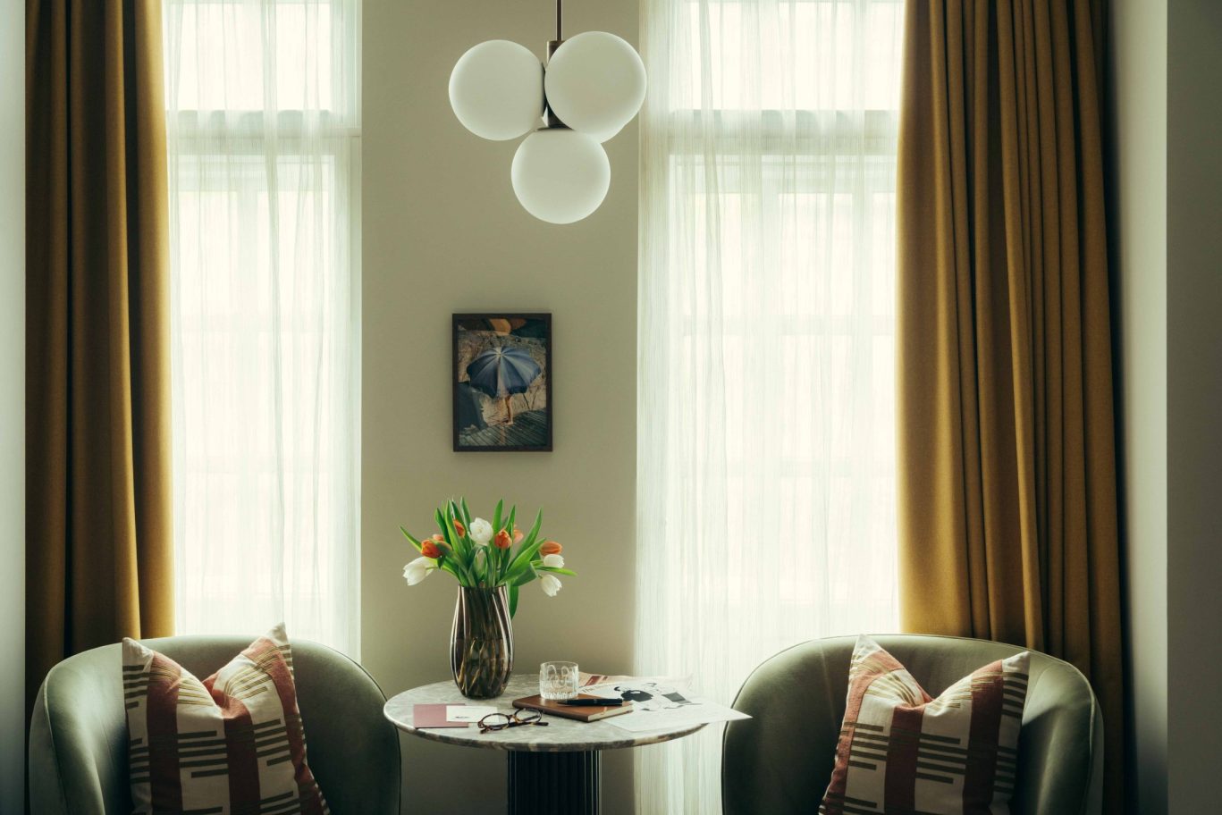 Cozy sitting area in Covent Garden with two armchairs, a small round table adorned with tulips, glasses, and papers, nestled between tall windows draped in gold curtains and illuminated by a modern globe light fixture above.