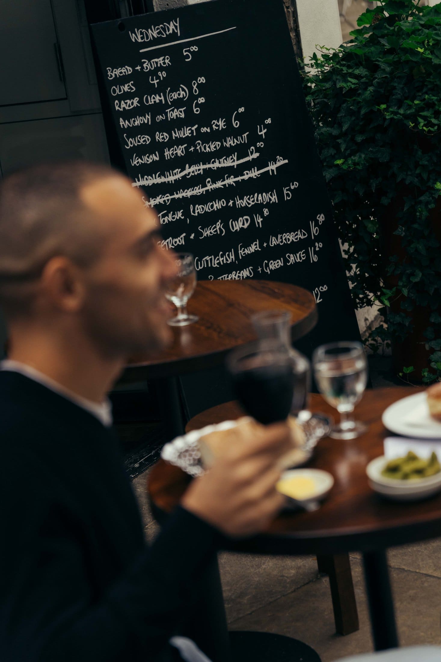 A man sits at an outdoor table with food and a glass of red wine. In the background, a chalkboard menu with handwritten dishes is visible next to green plants, evoking the charm of a Covent Garden cafe scene.