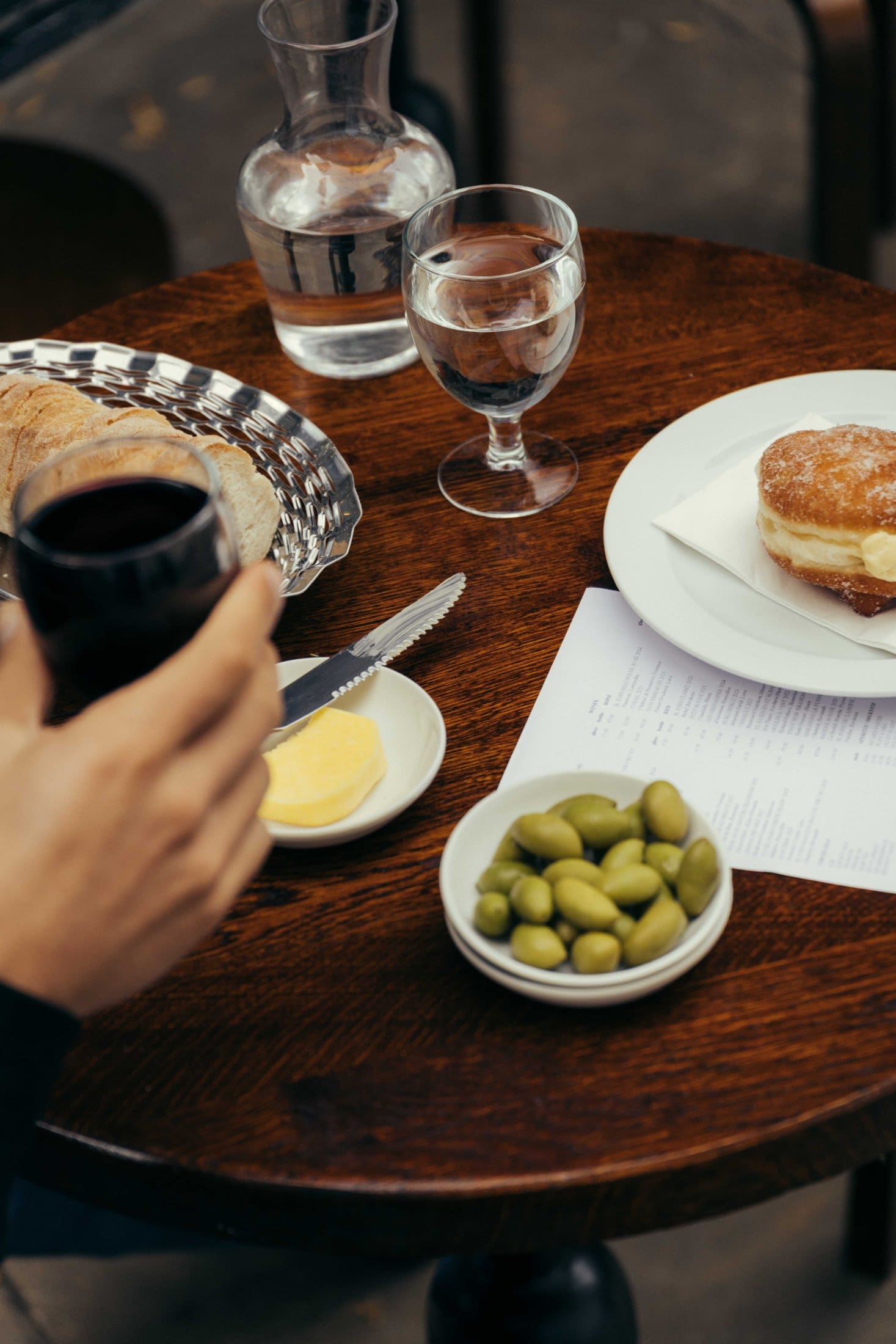 A hand holds a glass of red wine at a round wooden table in Covent Garden, set with bread, butter, green olives, a dessert pastry on a plate, a glass and carafe of water, and a menu.