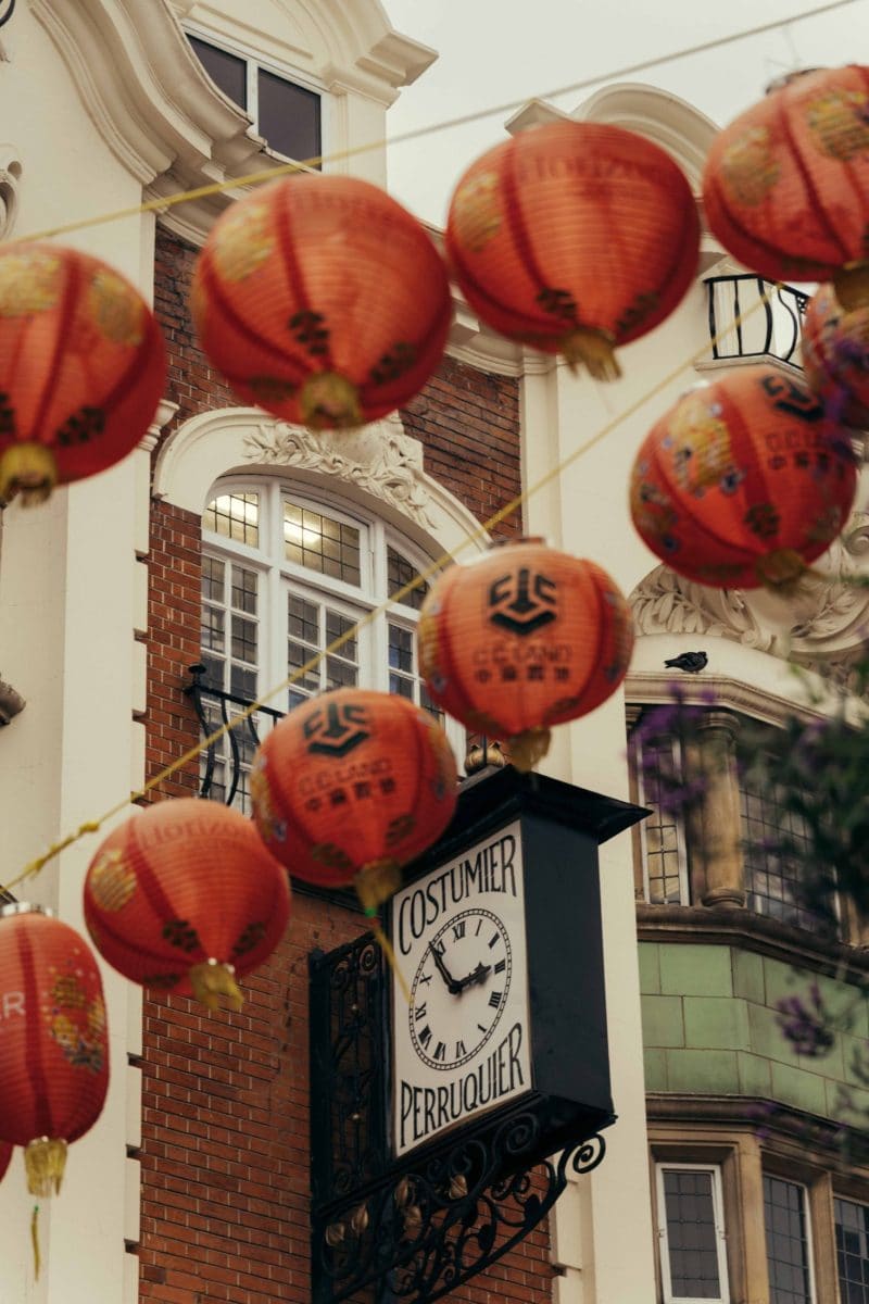 Red Chinese lanterns hang across a street in Covent Garden, in front of a brick building with large windows. A black and white clock sign below reads Costumier Perruquier, adding to the festive and lively scene.