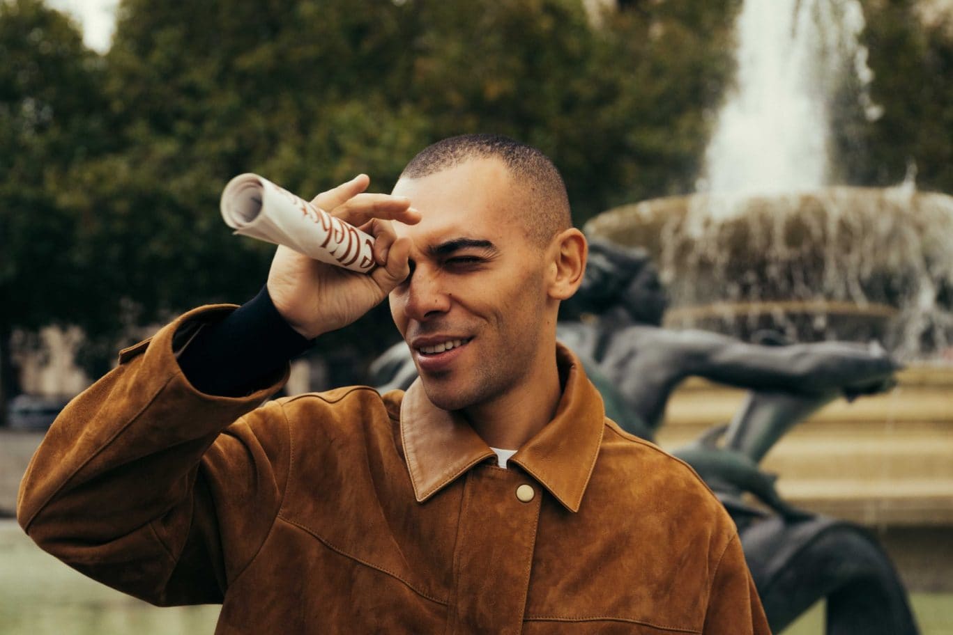 A man in a brown suede jacket playfully looks through a rolled-up paper as if it were a telescope, standing near the fountain and statue in Covent Garden, with trees framing the lively scene.