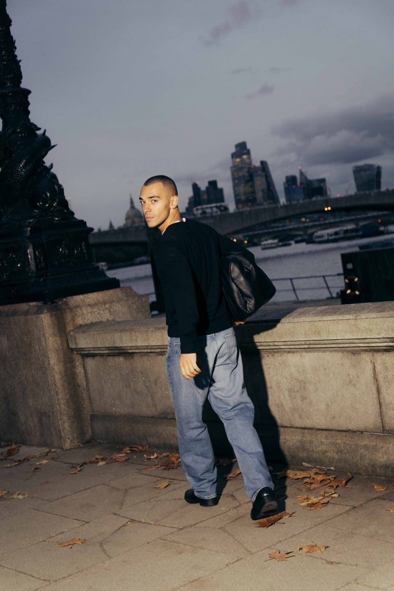 A person with short hair, wearing a black sweater and blue jeans, walks along a riverside path near Covent Garden at dusk, looking back over their shoulder. The city skyline and river are visible in the background.