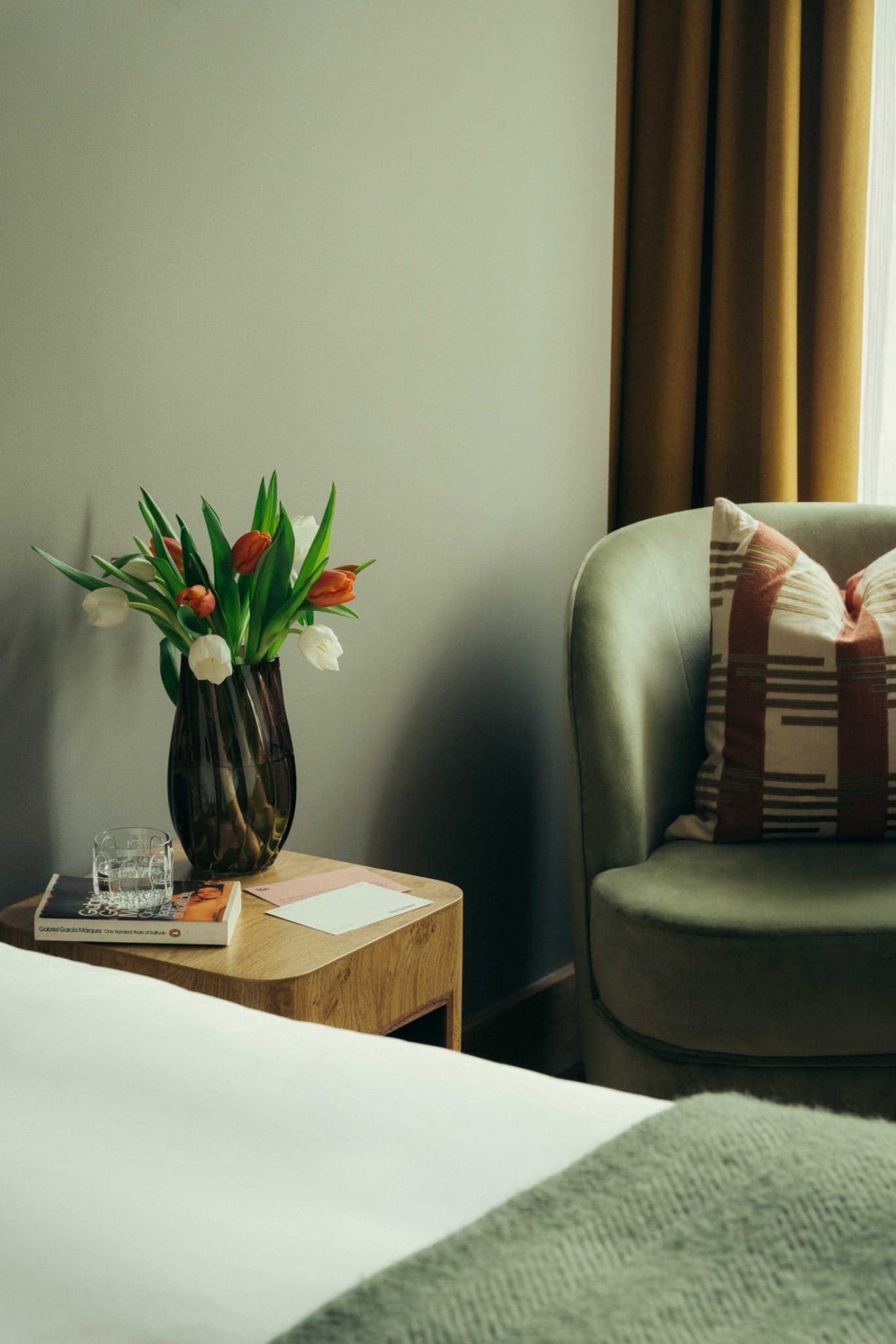A cozy corner reminiscent of Covent Garden, with a green armchair, patterned pillow, wooden side table holding tulips, a glass of water, magazines, and a note—nestled beside a bed draped in a green blanket.