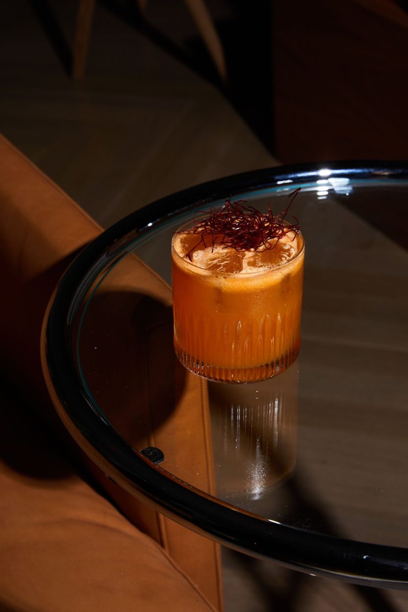A vibrant orange cocktail served in a glass with ice, garnished with fine red strands, sits on a glass-topped table next to a brown leather chair in a dimly lit Covent Garden setting.