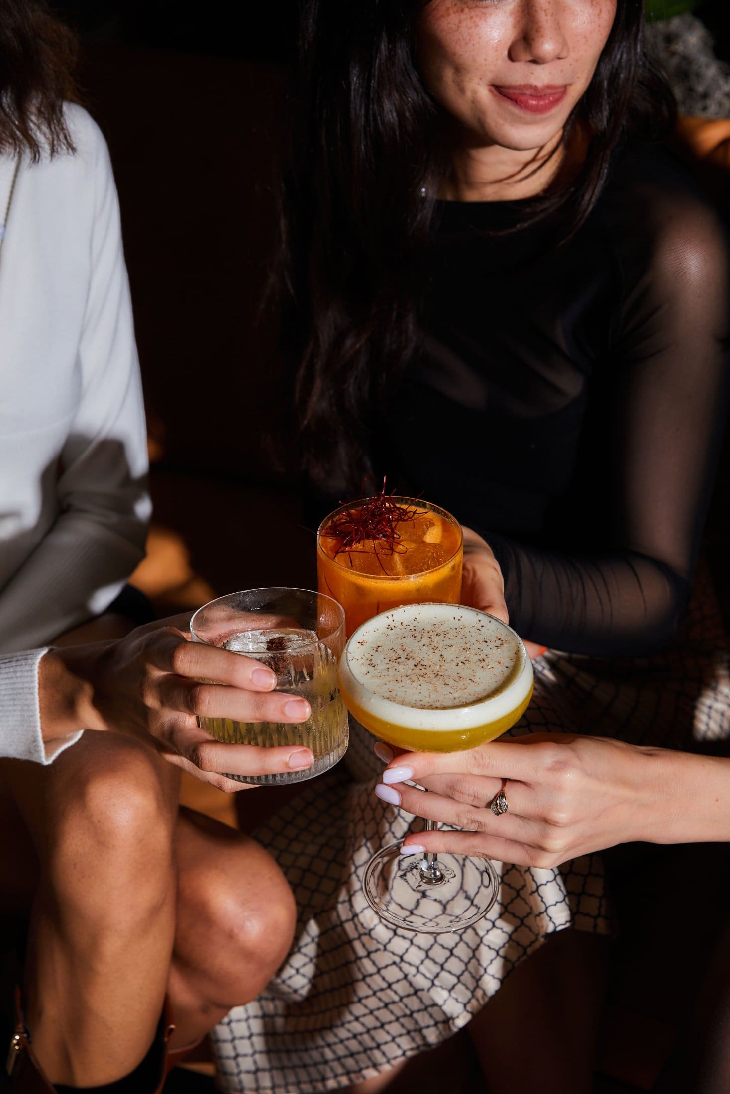 Two women sit close together, smiling and holding different cocktails in their hands. Sunlight highlights their arms and drinks, creating a warm, inviting Covent Garden atmosphere. Their faces are partially out of frame.